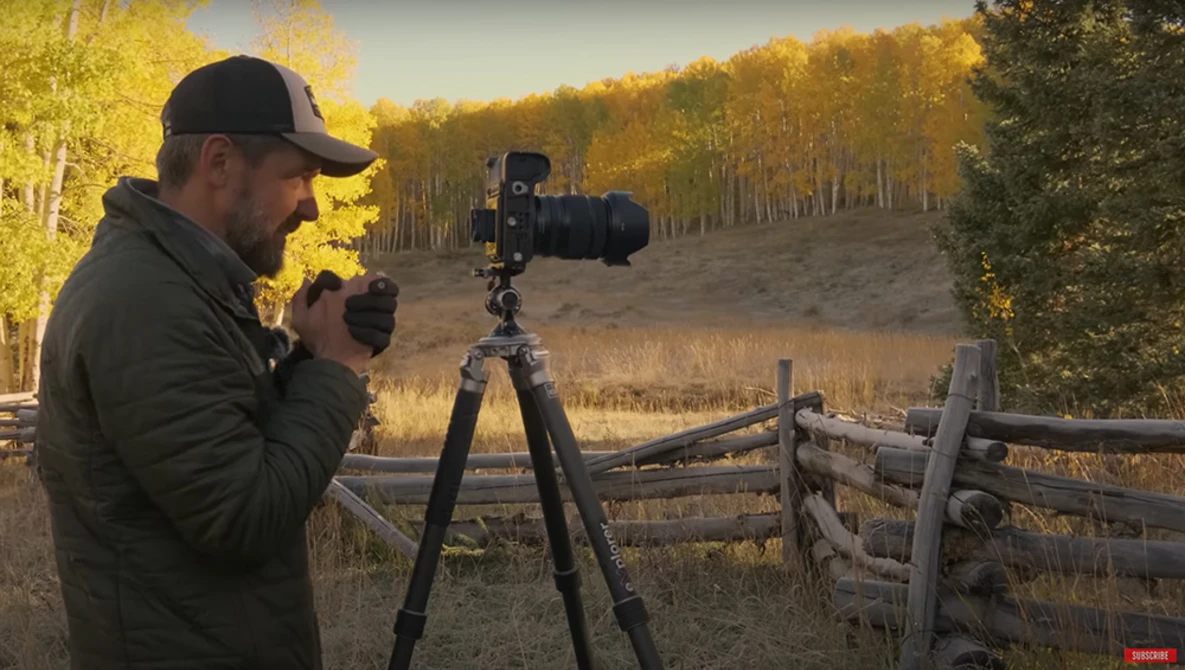 Photographer with camera on tripod composing a landscape shot during golden hour near a wooden fence.