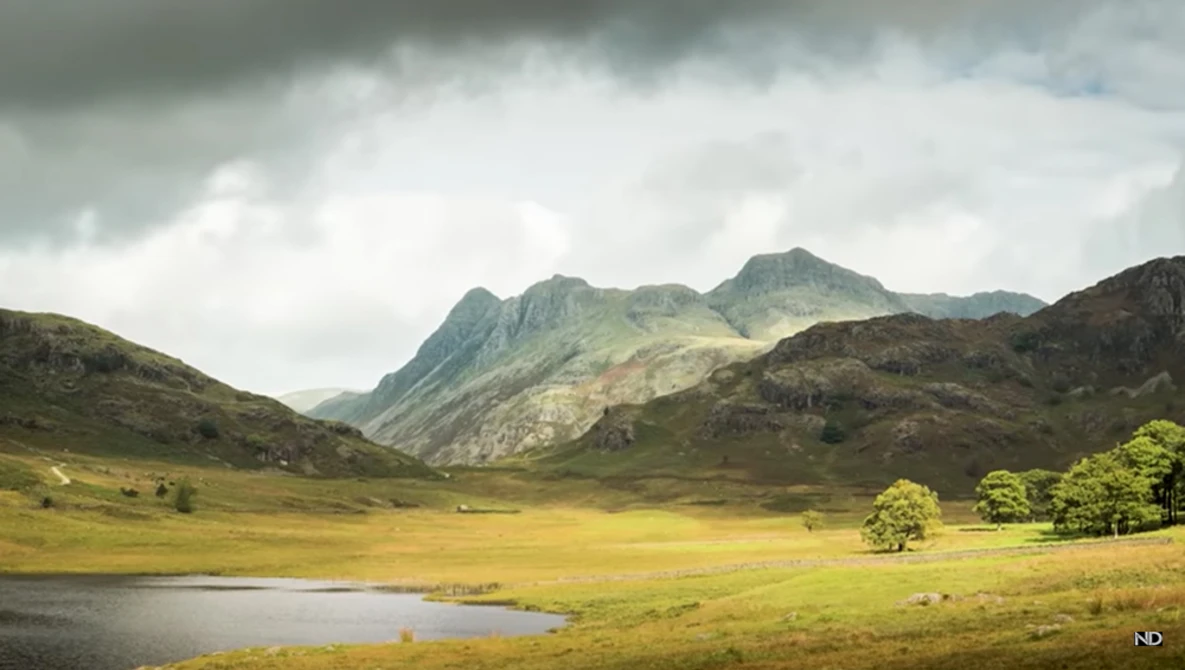 Mountain valley landscape with dramatic peaks, green meadows, and a small lake under stormy skies.
