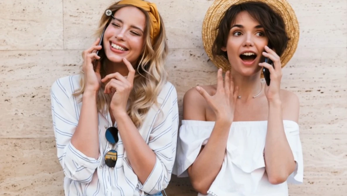 Two women wearing straw hats and white tops smiling while talking on mobile phones against a brick wall.