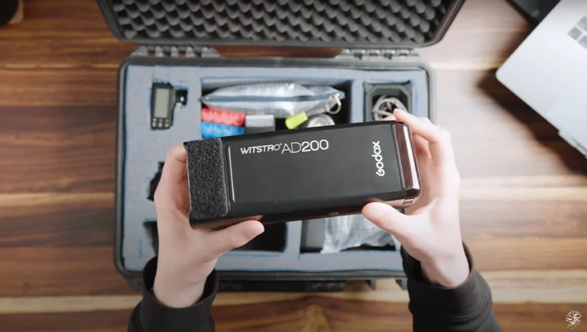 Hands holding a Godox Wistro AD200 flash unit above an open equipment case on a wooden surface.