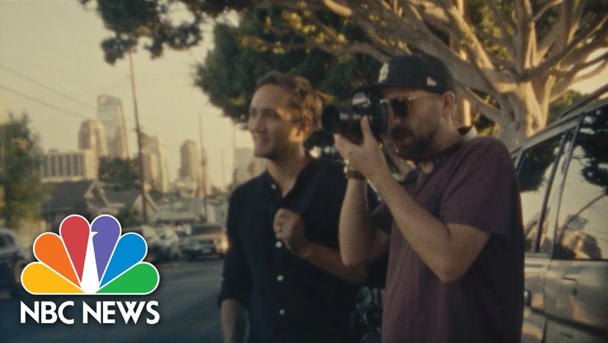 Cameraman filming two men on an urban street with city skyline in background during golden hour.