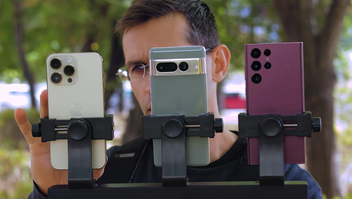 Three smartphones in different colors held by a person wearing glasses, each mounted on a black device holder for comparison.