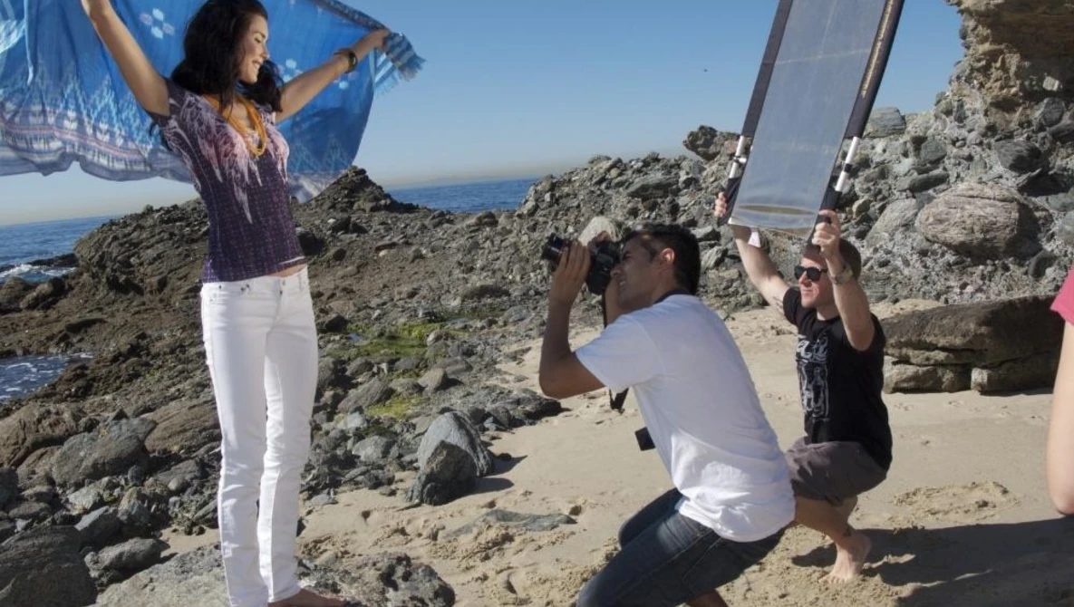 Photographer crouching to capture a portrait of a woman posing on a rocky coastal outcrop with ocean and sky in background.