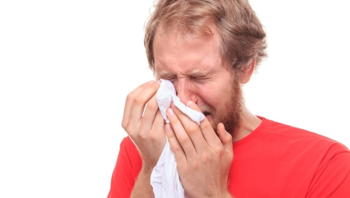Man in red shirt holding tissue to his nose while sneezing or blowing his nose.