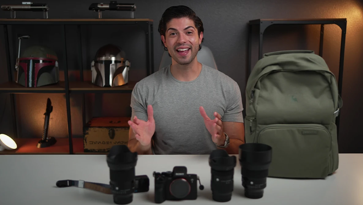 Photographer with camera equipment and lenses arranged on desk, shelving with gear visible behind.