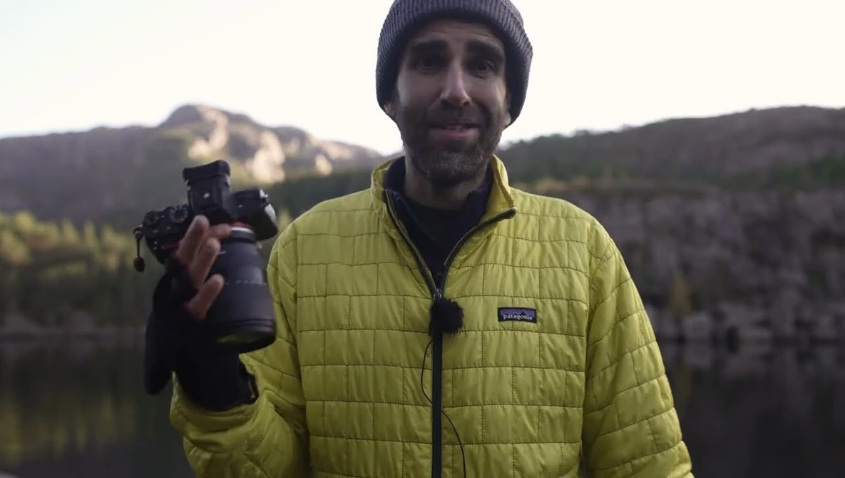 Photographer holding a telephoto lens while smiling at the camera with forested mountains and a lake visible behind him.