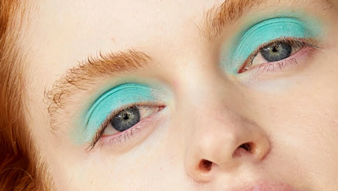 Close-up portrait of a person with turquoise eyeshadow and reddish-brown hair against a neutral background.