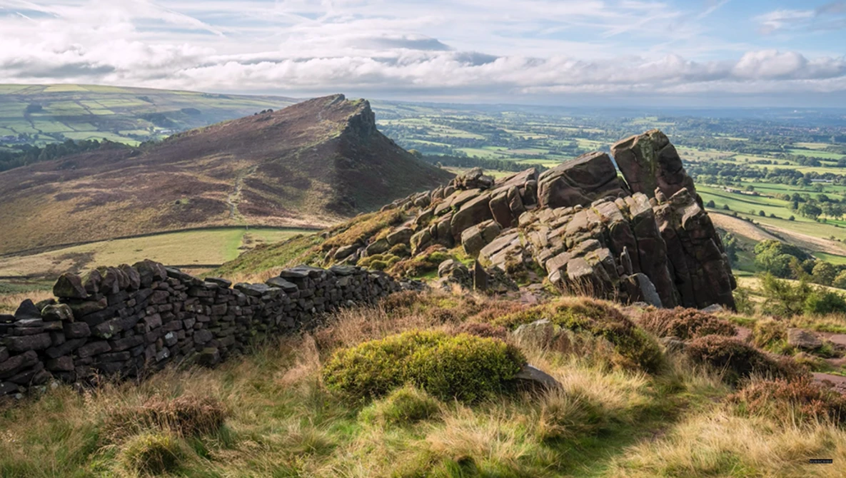 Rocky hilltop outcrop in foreground with moorland and distant peaked mountain under dramatic cloudy sky.