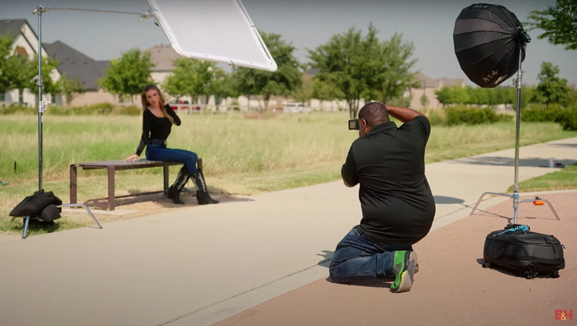 Portrait photographer crouching with camera on outdoor concrete set, shooting subject seated on bench with reflector and key light positioned nearby.