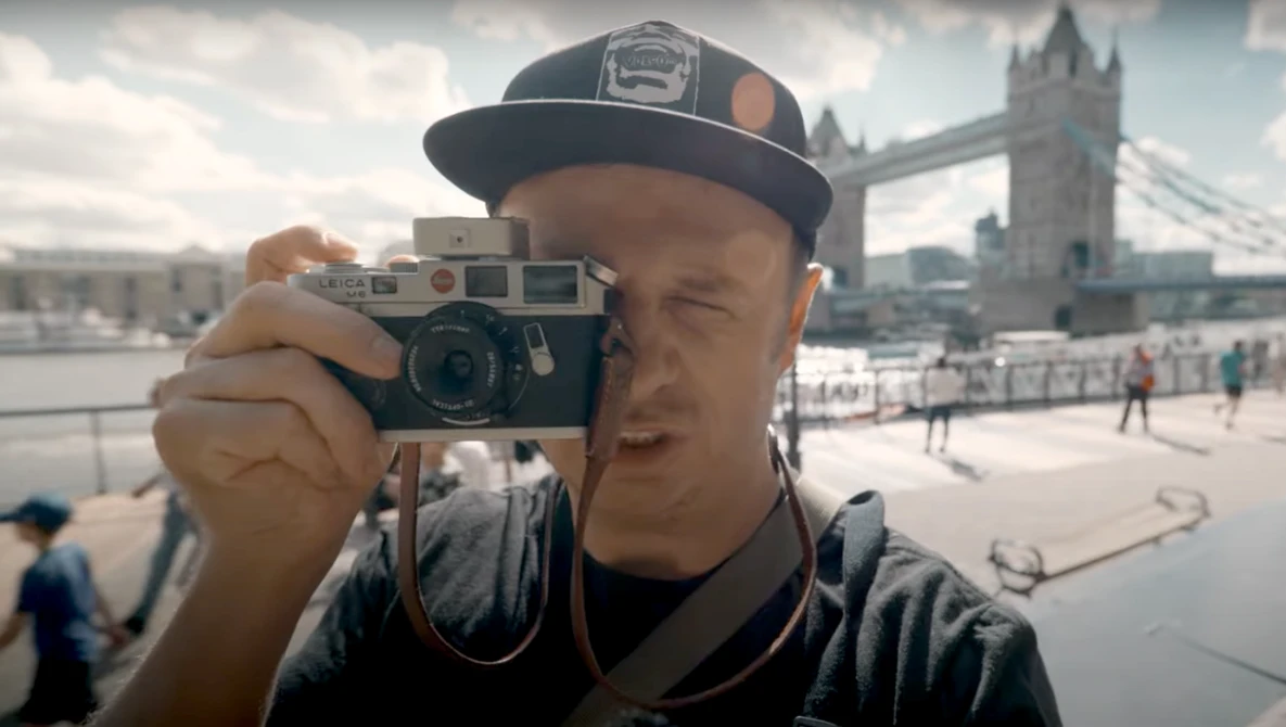 Photographer holding a vintage rangefinder camera up to his face, with Tower Bridge visible in the background.
