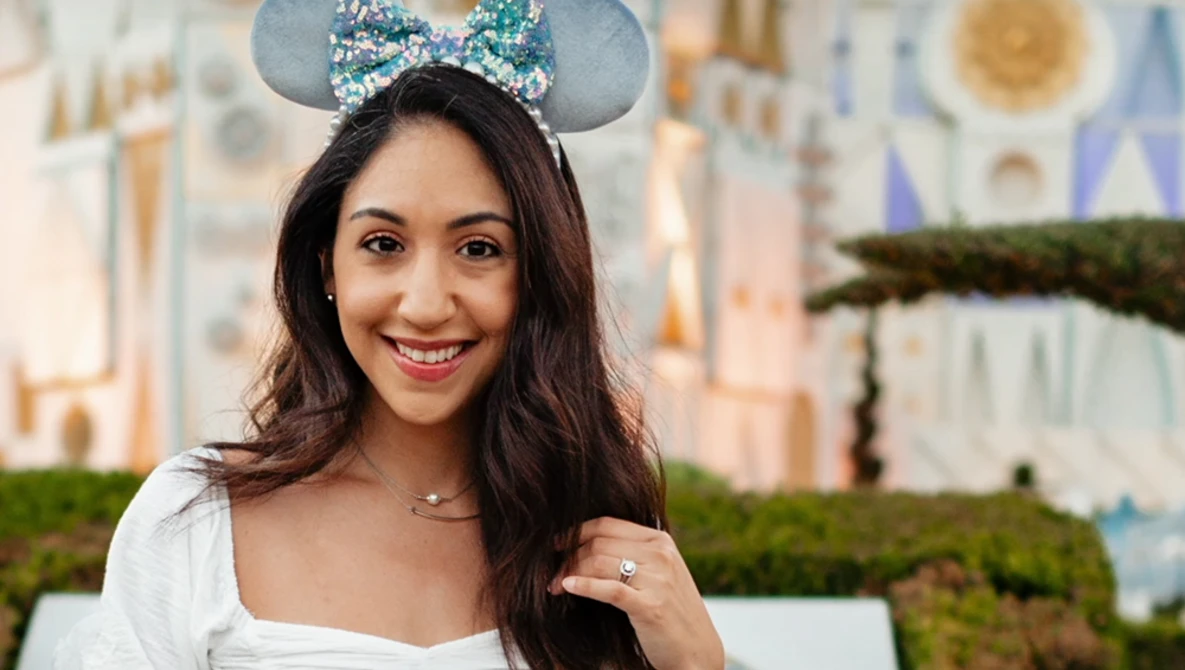 Woman wearing a sparkly blue Mickey ear headband smiles at the camera in front of a castle.