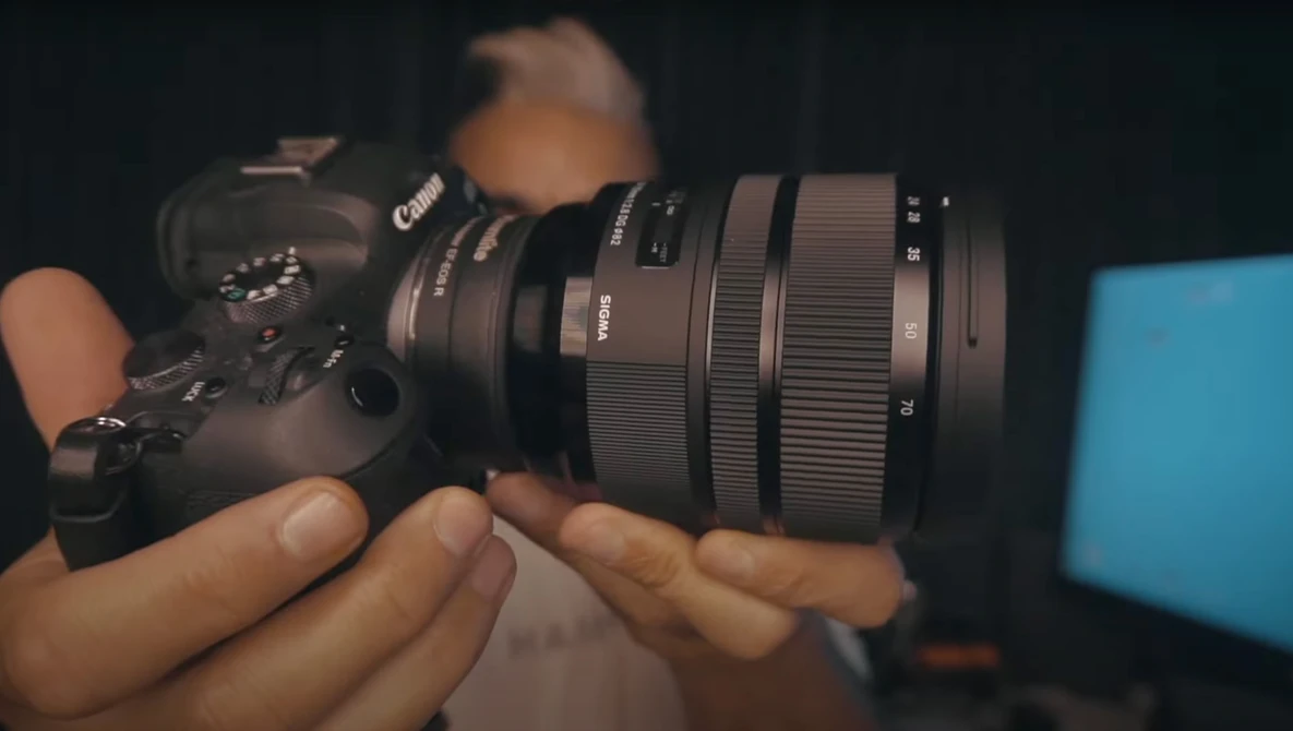 Hands holding a Canon camera with a telephoto lens attached, shown in close-up against a dark background.