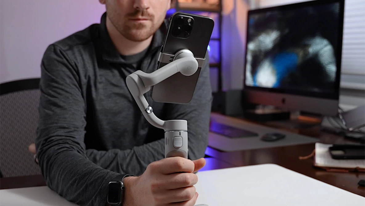 Man holding a three-axis gimbal stabilizer with a smartphone mounted on top in a home studio setting.