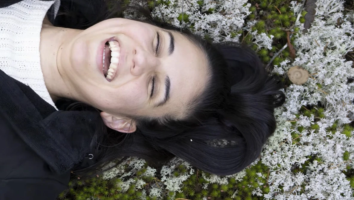 Person with dark hair and bright smile lying on lichen-covered ground, shot from above.