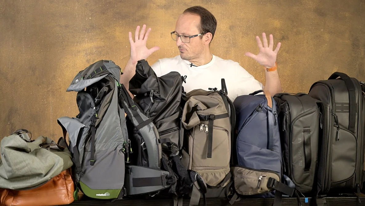 Man surrounded by camera backpacks and travel gear, hands raised in a gesture of overwhelm or indecision.