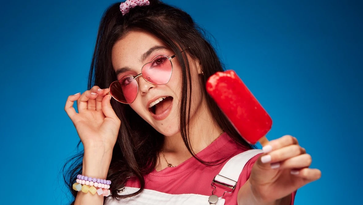 Young woman wearing pink heart-shaped sunglasses and a red and white shirt against a blue background.