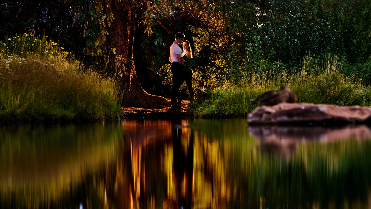 Couple embracing by a riverside at golden hour, surrounded by tall grass and trees with warm sunlight reflecting on calm water.