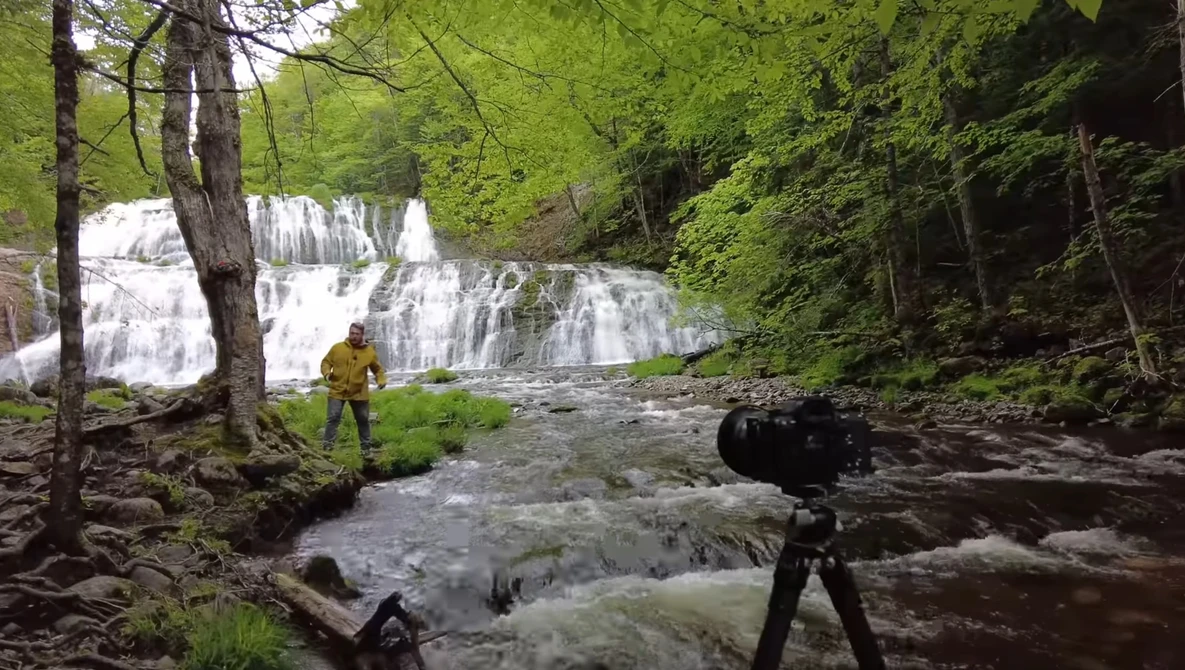 Photographer with camera on tripod capturing a multi-tiered waterfall in a lush forest stream.