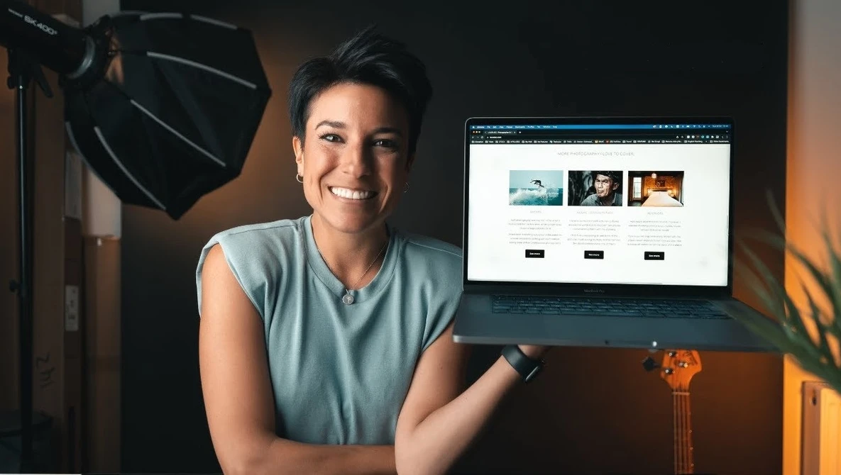 Creative professional smiling at camera while pointing to website portfolio displayed on monitor in studio setting.