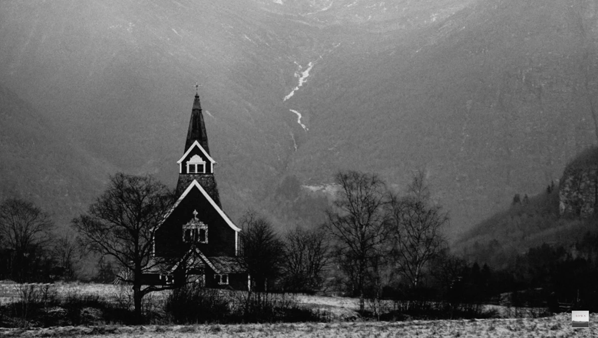 Wooden church with steeple in snowy valley beneath mountains and waterfall.