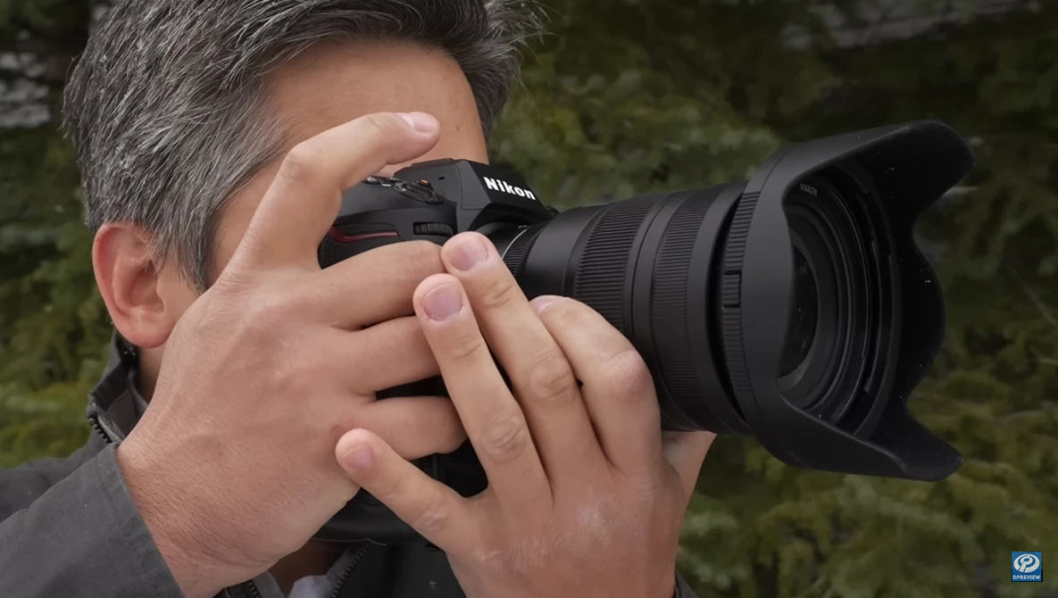 Photographer holding a Nikon DSLR camera with telephoto lens to his eye while composing a shot outdoors.