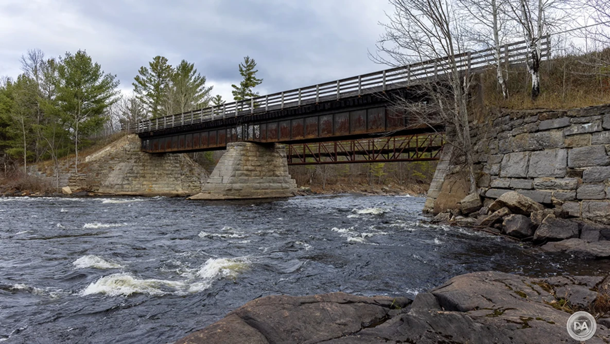 Steel truss bridge spanning a rocky river with bare trees and overcast sky in the background.