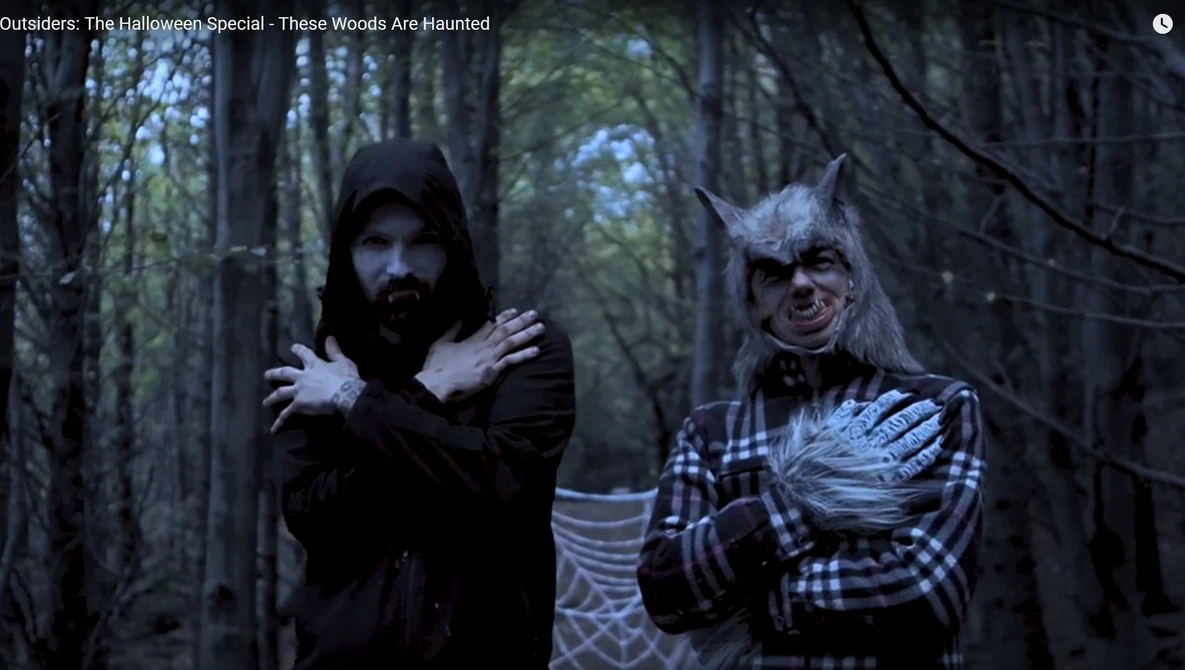 Two costumed characters in a dark forest setting, one in black robes and the other in plaid clothing with theatrical makeup.