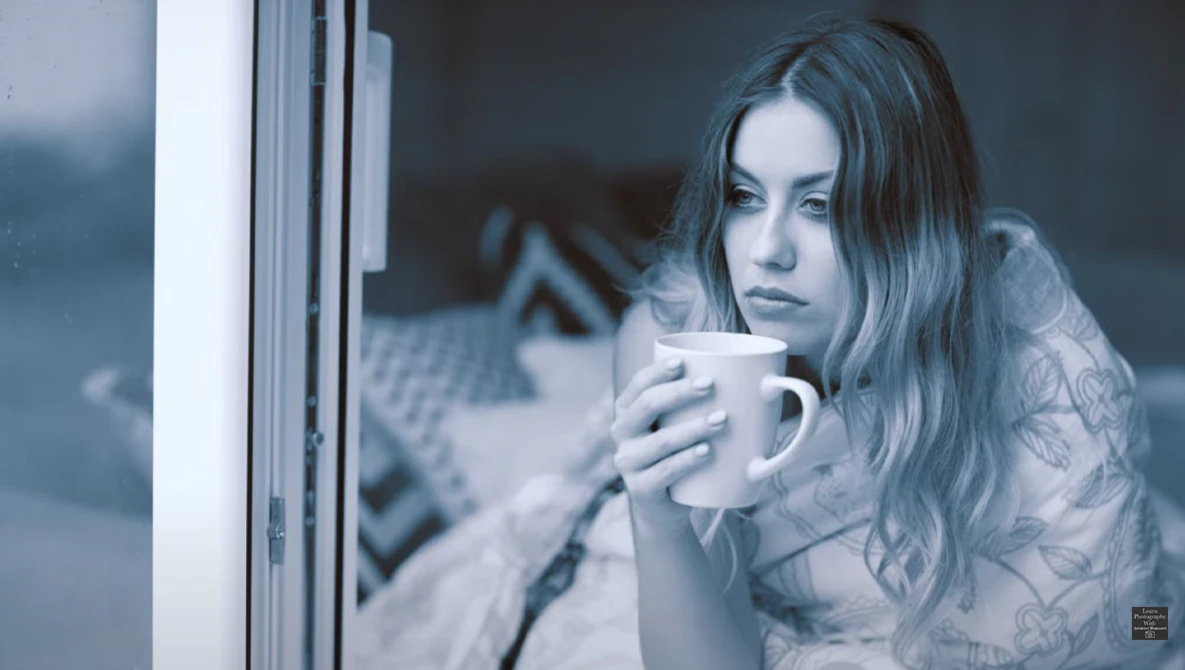 Woman looking out a window while holding a mug, shot through glass with cool blue color grading.