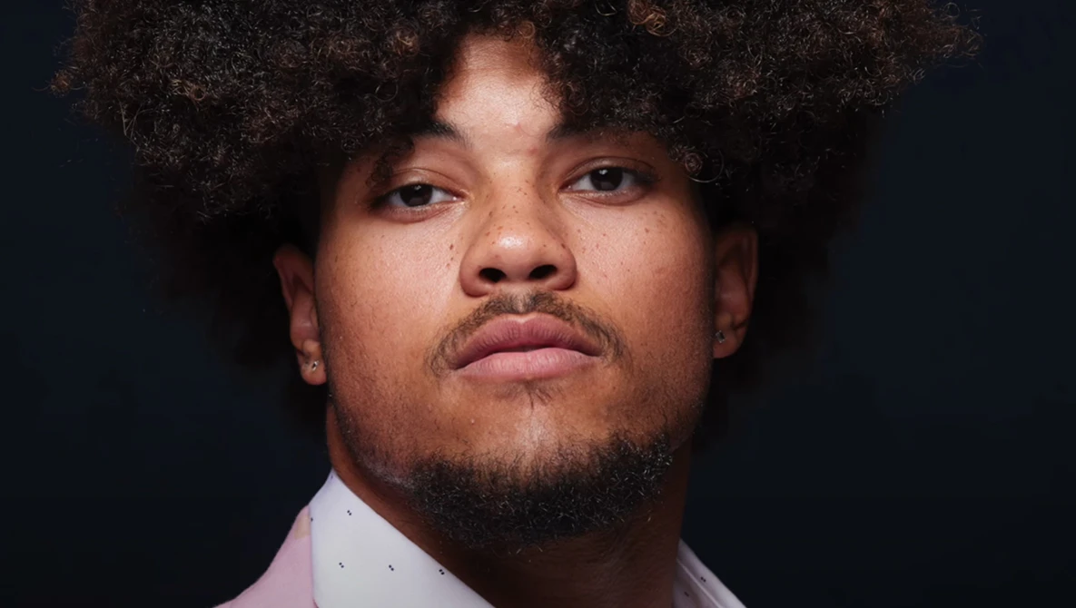 Portrait of a man with textured hair against a dark background, wearing a light pink shirt.