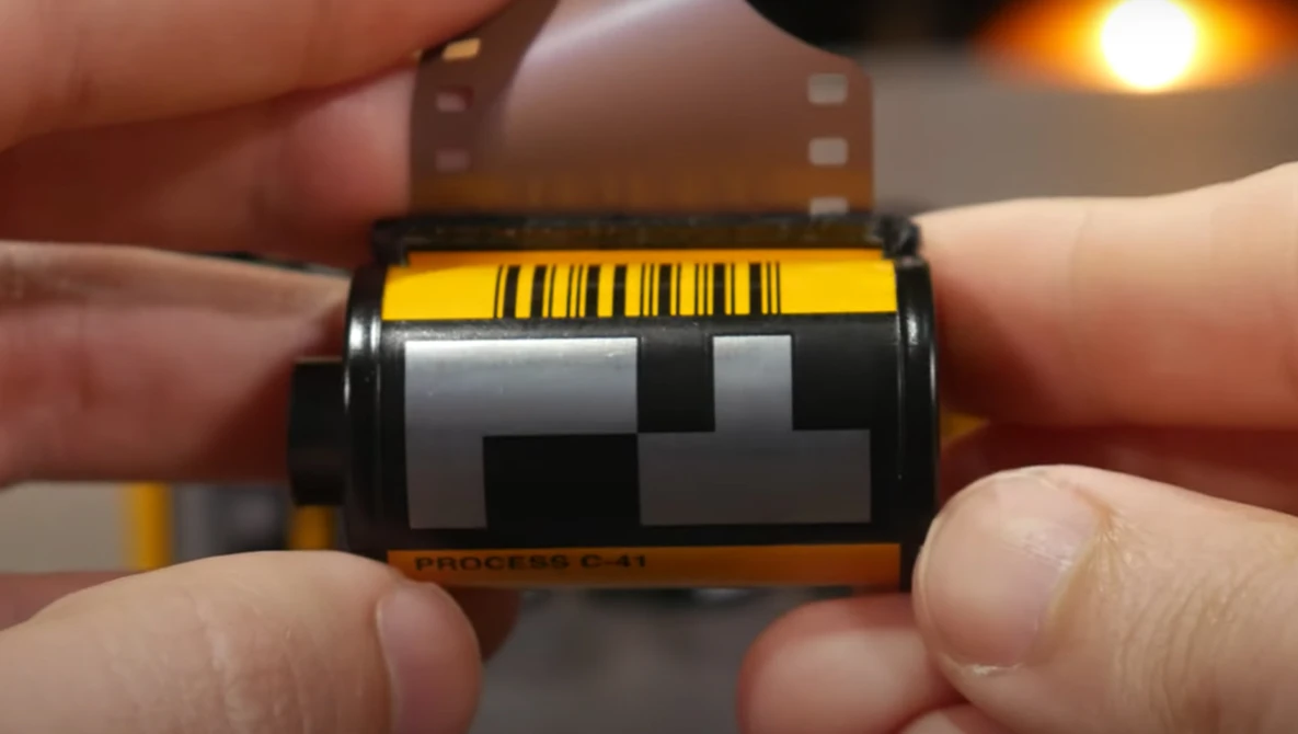 Close-up of a film canister being held in hands, showing the yellow barcode label and gray leader tape.