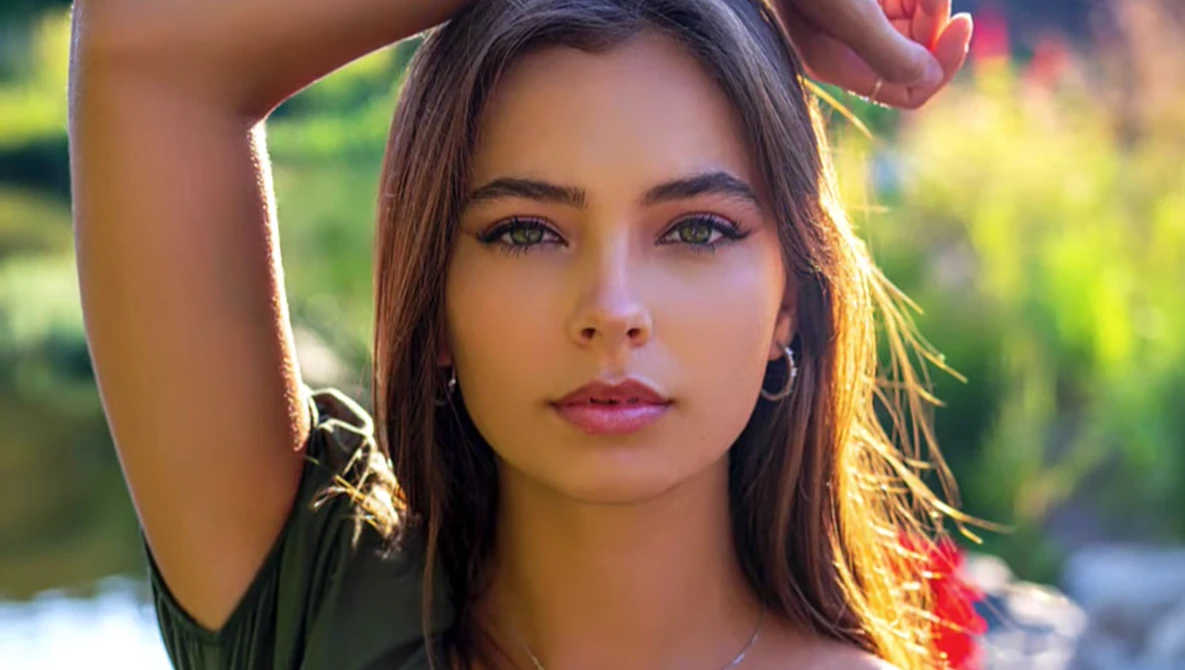 Portrait of a young woman with long dark hair in golden hour sunlight, hand raised to her head, looking directly at camera.