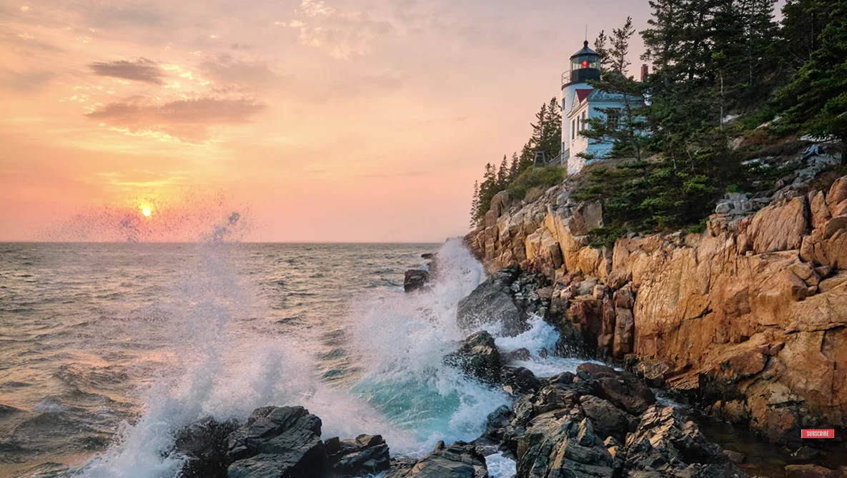 Crashing waves against rocky coastline at sunrise with lighthouse on forested cliff.