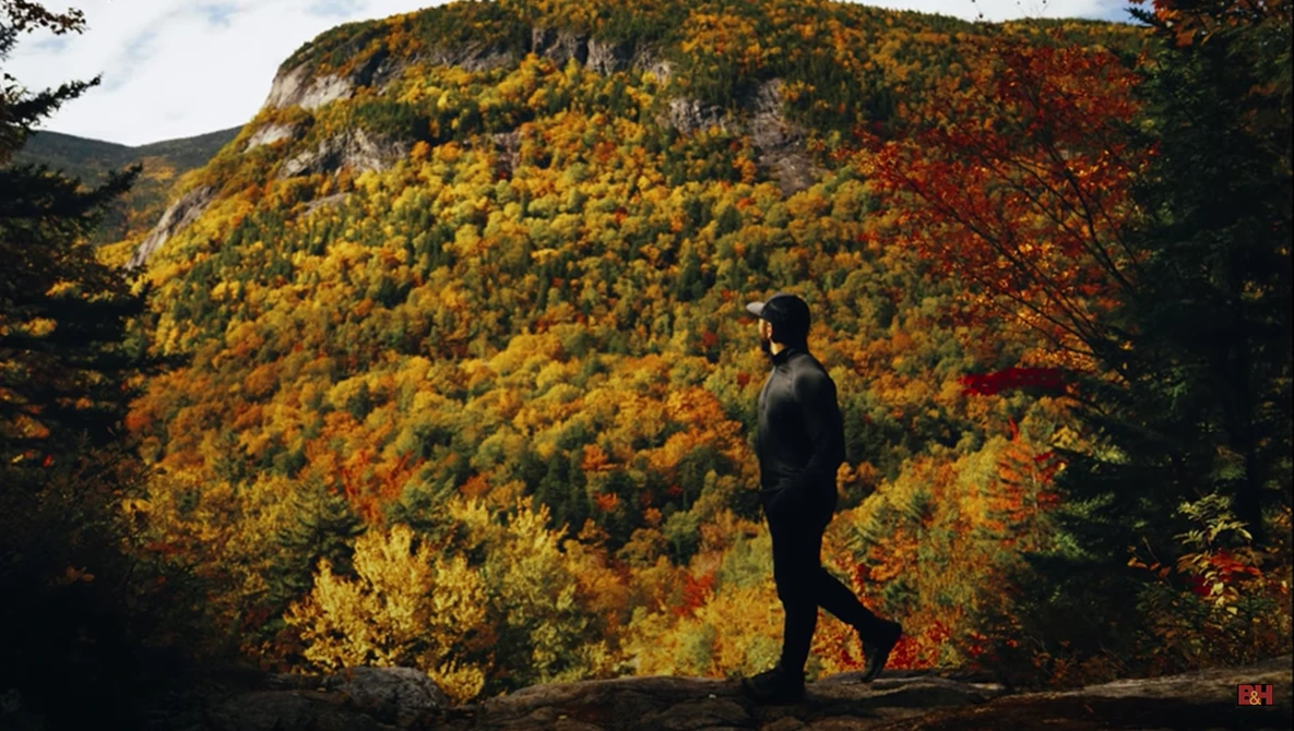 Person standing on a rocky outcrop facing a mountainside covered in vibrant autumn foliage.