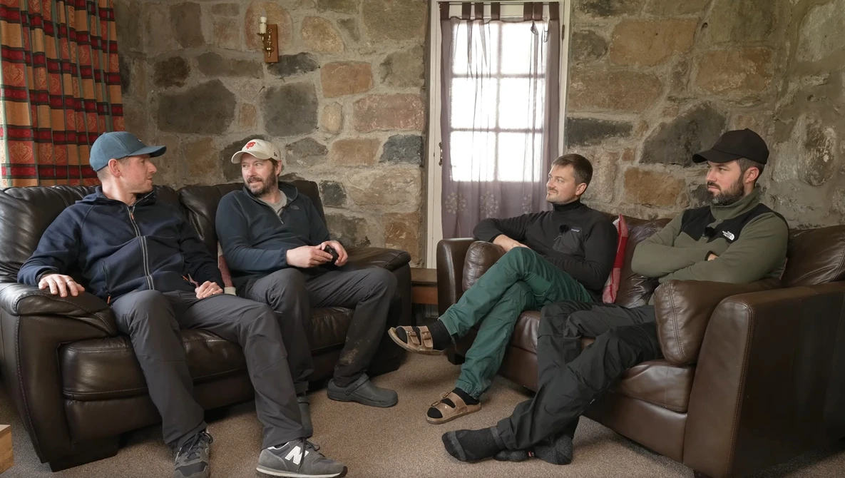 Four men sitting on leather couches in a stone-walled room engaged in conversation.