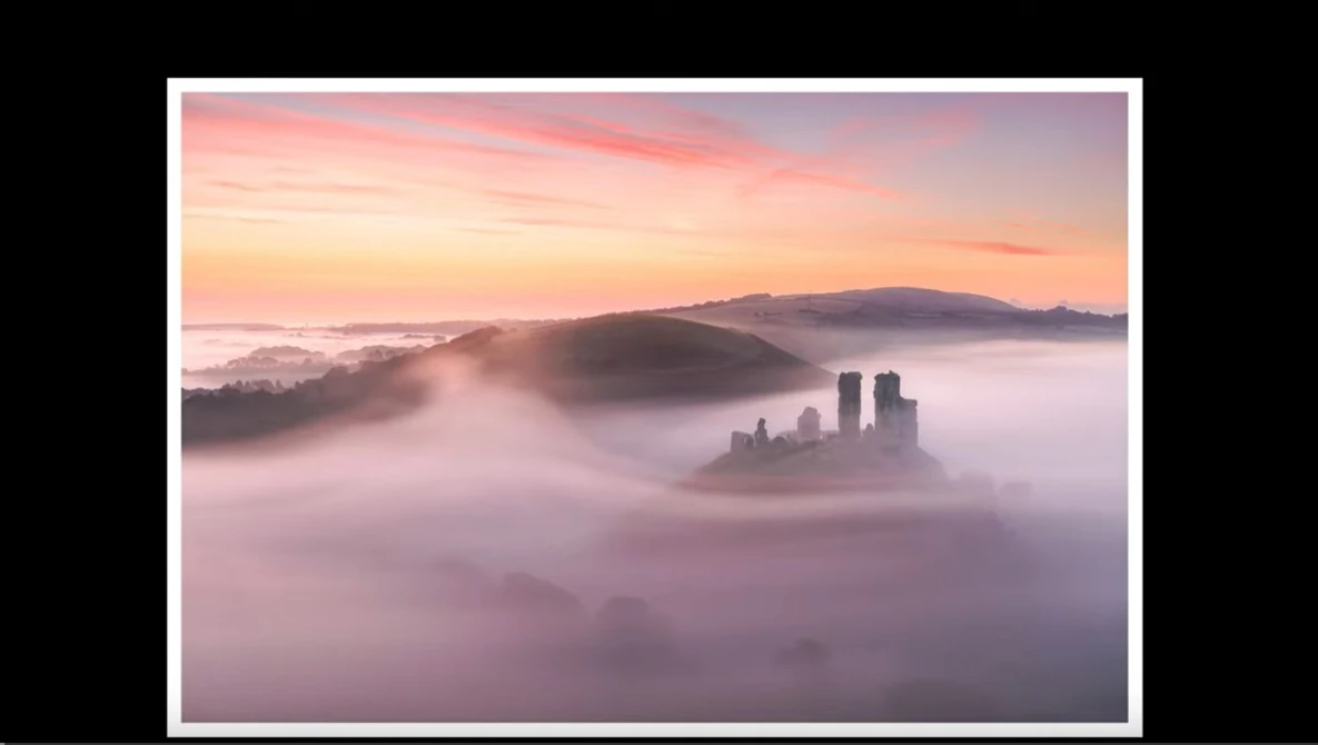 Medieval castle ruins emerging from thick fog at sunrise, with layered mountains visible in the distance.