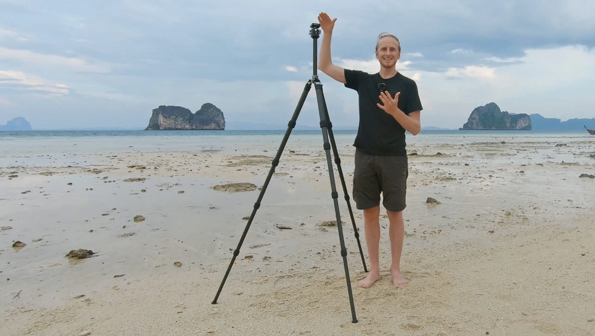 Photographer with tripod on beach gestures toward camera during golden hour shoot.