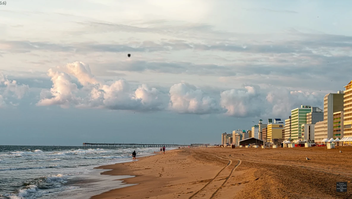 Beach scene at sunset with high-rise hotels lining the shore, a wooden pier extending into calm waters, and dramatic cloud formations on the horizon.