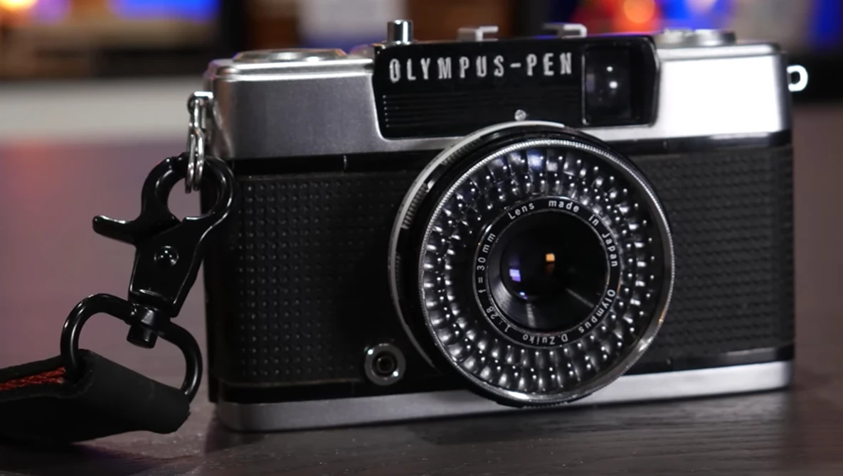 Olympus PEN half-frame camera with rangefinder lens and wrist strap, displayed on dark surface with blurred background lights.