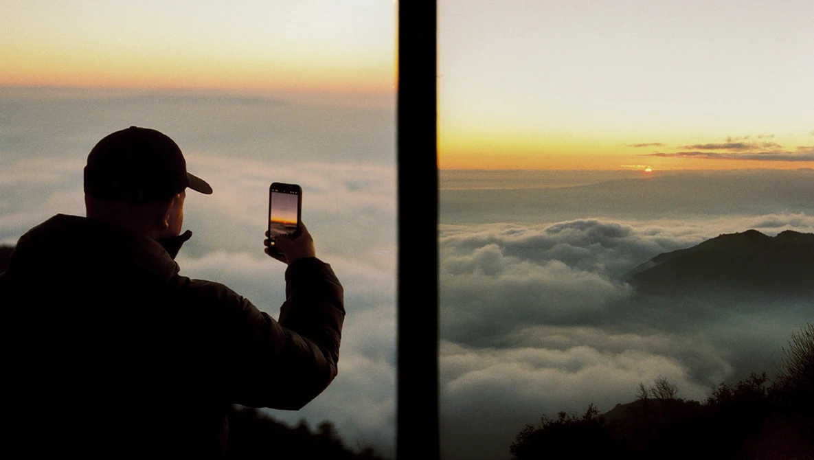 Silhouetted figure photographing a sunrise above clouds with a smartphone.