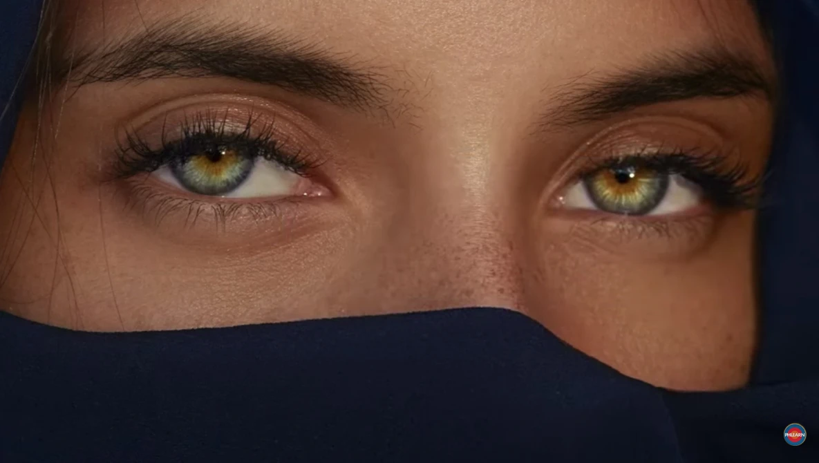 Close-up portrait of a person's eyes with golden-amber irises and dark brows, lower face obscured by dark fabric.