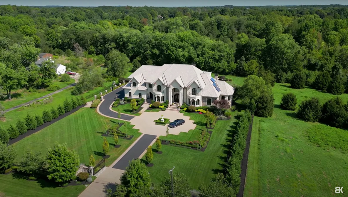 Aerial view of a large two-story stone and brick residence with curved driveway on manicured grounds surrounded by mature trees.