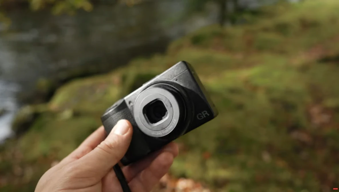 Hand holding a compact digital camera with a black body and silver lens ring outdoors.