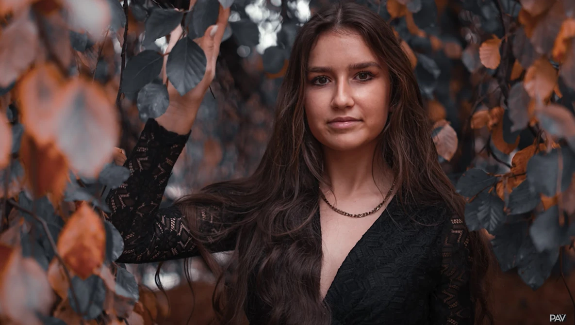 Portrait of a woman with long dark hair framed by autumn foliage in warm orange and brown tones.