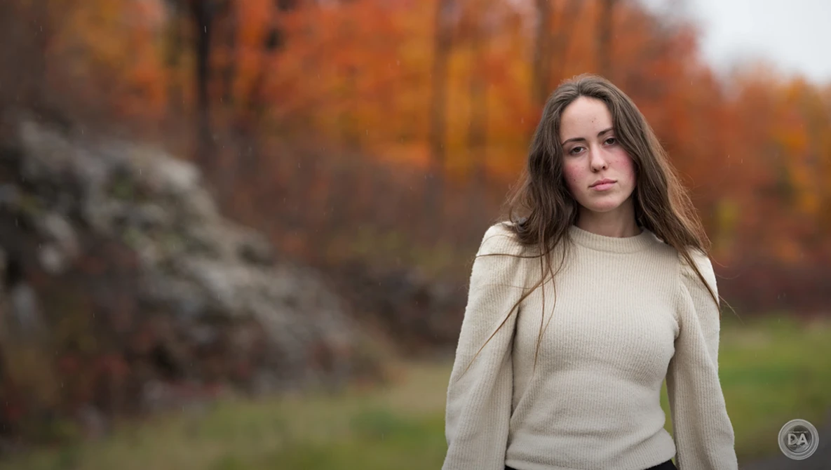 Woman in cream sweater standing outdoors with autumn foliage in soft focus background.