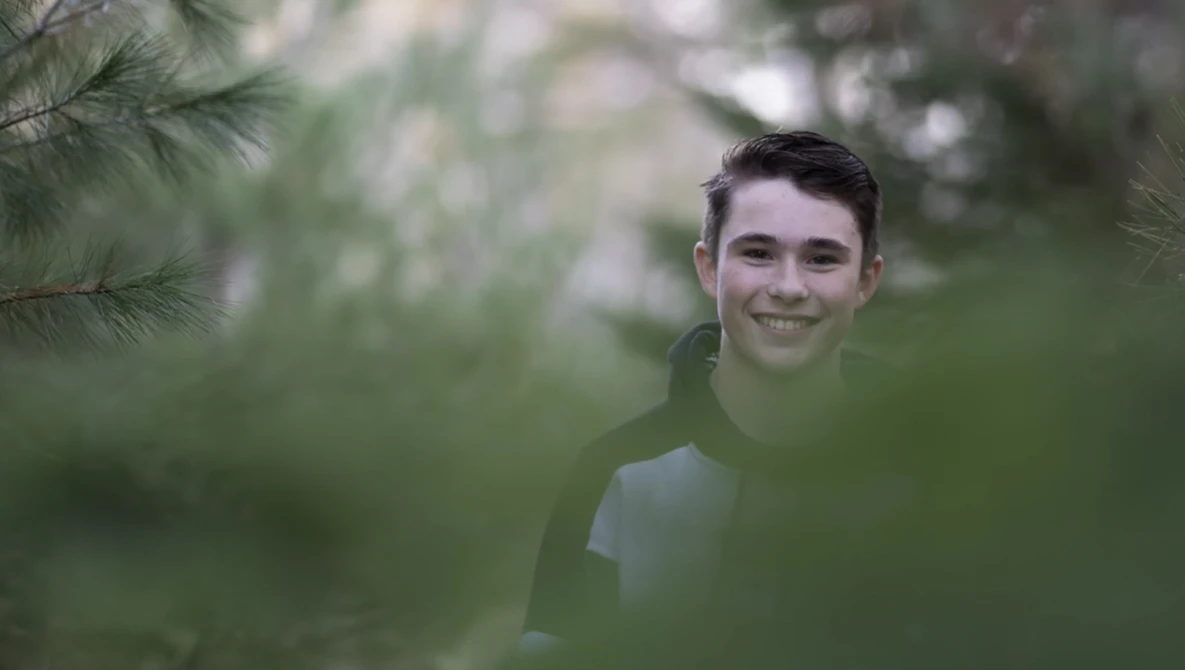 Young man smiling at camera surrounded by soft green foliage and evergreen branches.