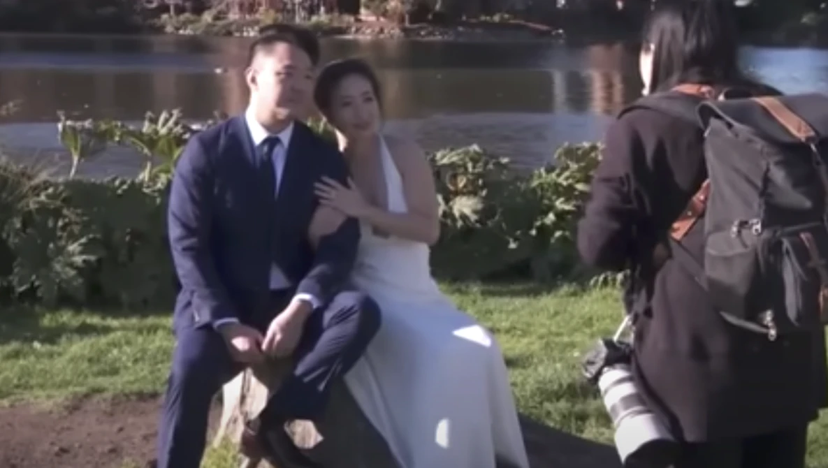 Wedding photographer shooting bride and groom posing by waterside during outdoor ceremony.