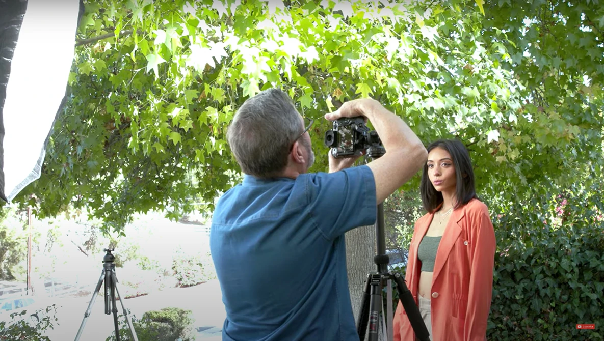 Photographer using a DSLR camera on a tripod to capture a portrait of a woman in an orange cardigan under leafy green canopy.