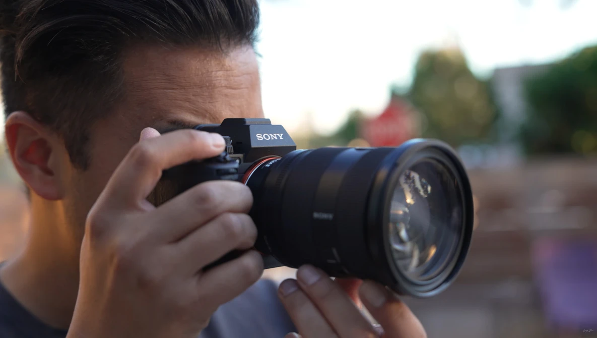 Photographer holding Sony mirrorless camera with telephoto lens, shooting in outdoor setting with blurred background.