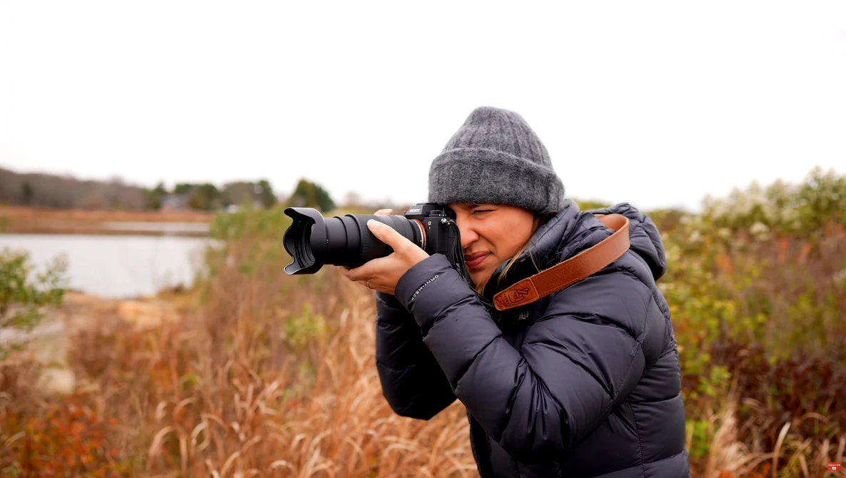 Photographer in winter gear holding a DSLR with telephoto lens while shooting outdoors in a rural landscape.