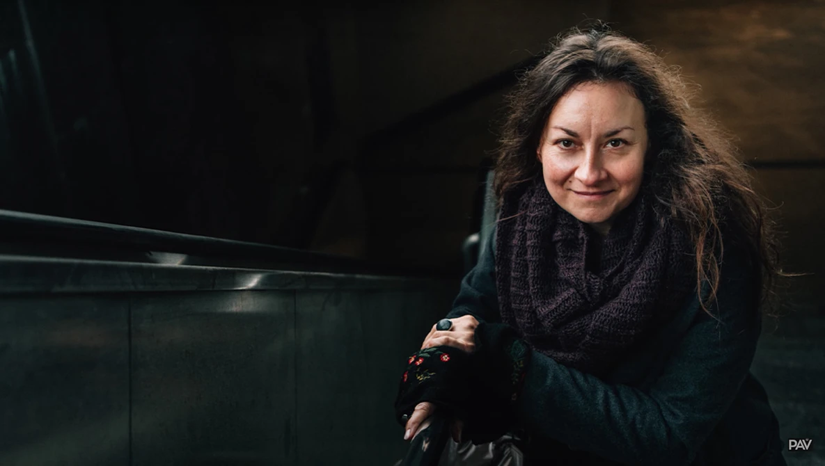 Woman with long dark hair wearing a black scarf and jacket, smiling at the camera in an indoor setting.